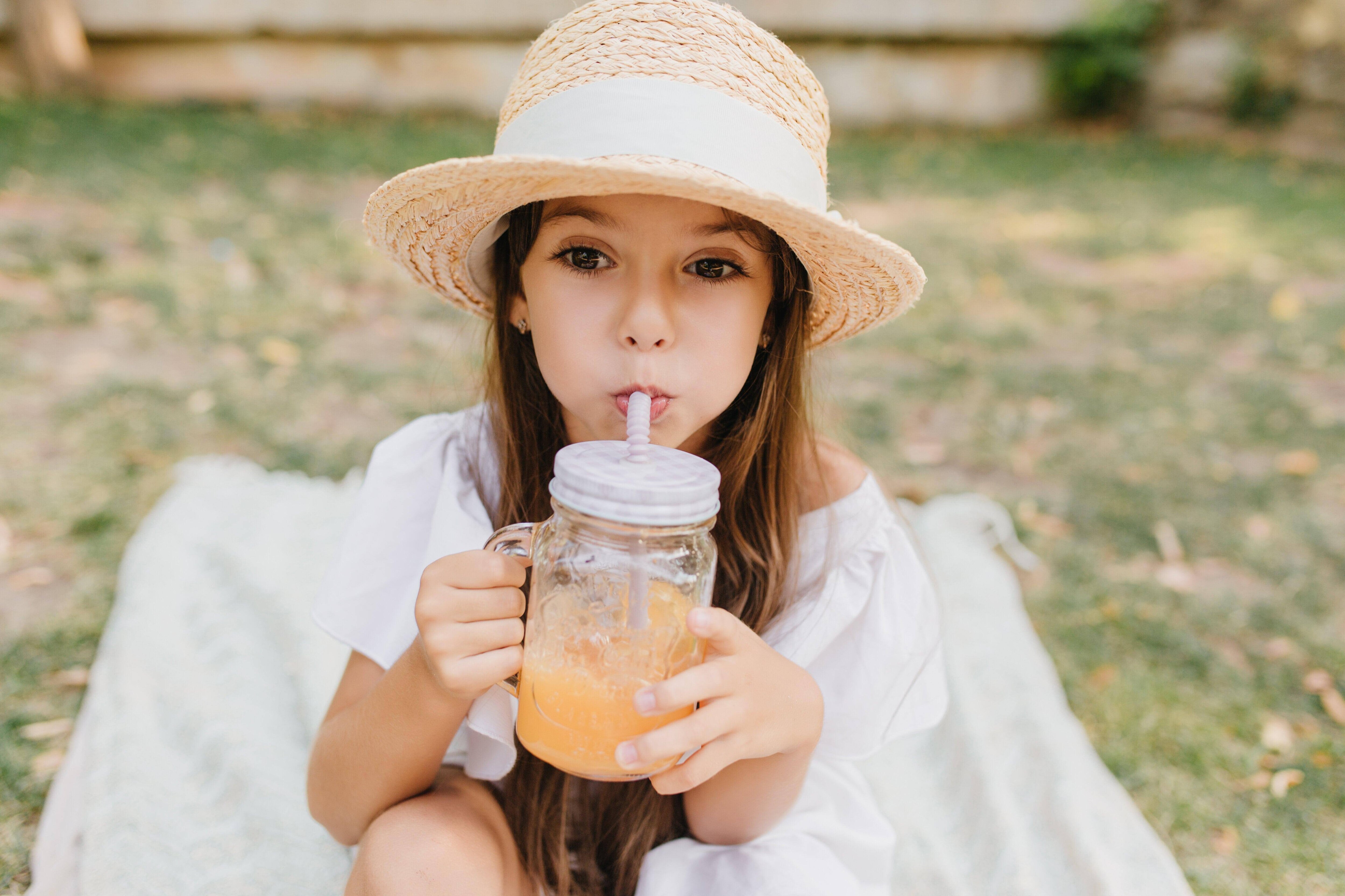 Bambina con cappello di paglia beve frullato di frutta da bicchiere di vetro