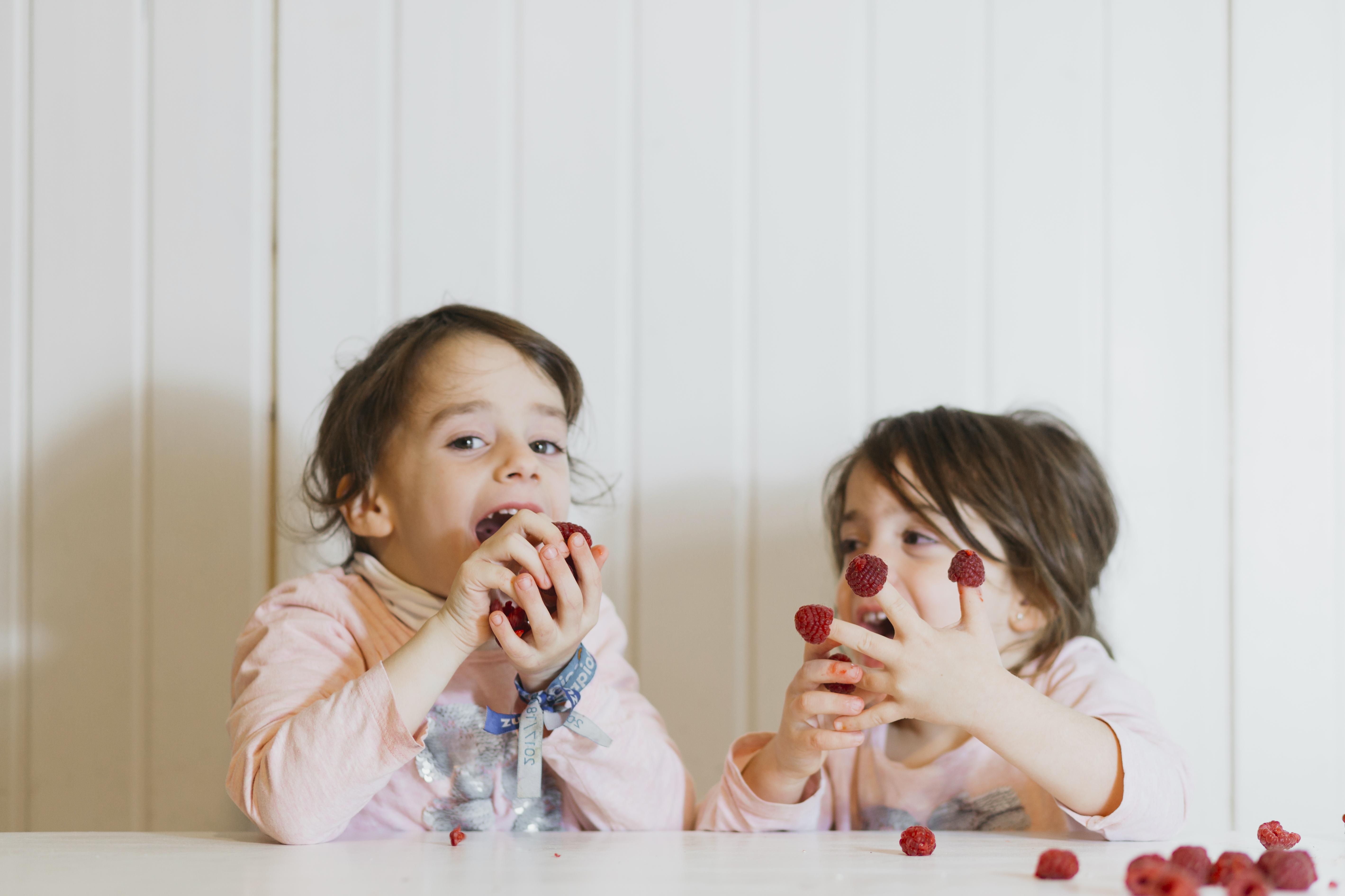 Due bambini a tavola giocano e mangiano lamponi rossi su sfondo bianco