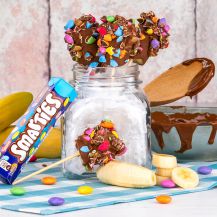 Barattolo con SMARTIES® Banana Sticks su tavolo con accanto tubetto di smarties, banane e ciotola di cioccolato con cucchiaio di legno