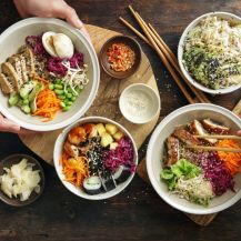 Mani che tengono piatto con verdure, legumi e sushi al tonno sul tavolo, mano con bacchette di legno e piatti di verdure e legumi 
