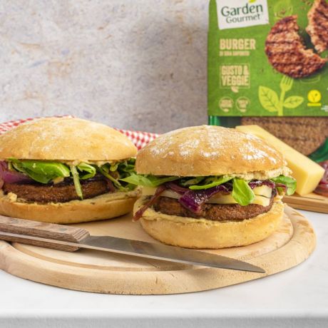 Scopri la ricetta del Burger con cipolle caramellate 2 Burger con cipolle caramellate su tavola di legno e confezione Garden Gourmet sullo sfondo